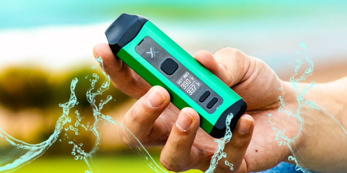 A hand holding a green Exxus Mini Plus with water splashes around it, set against a blurred outdoor background.